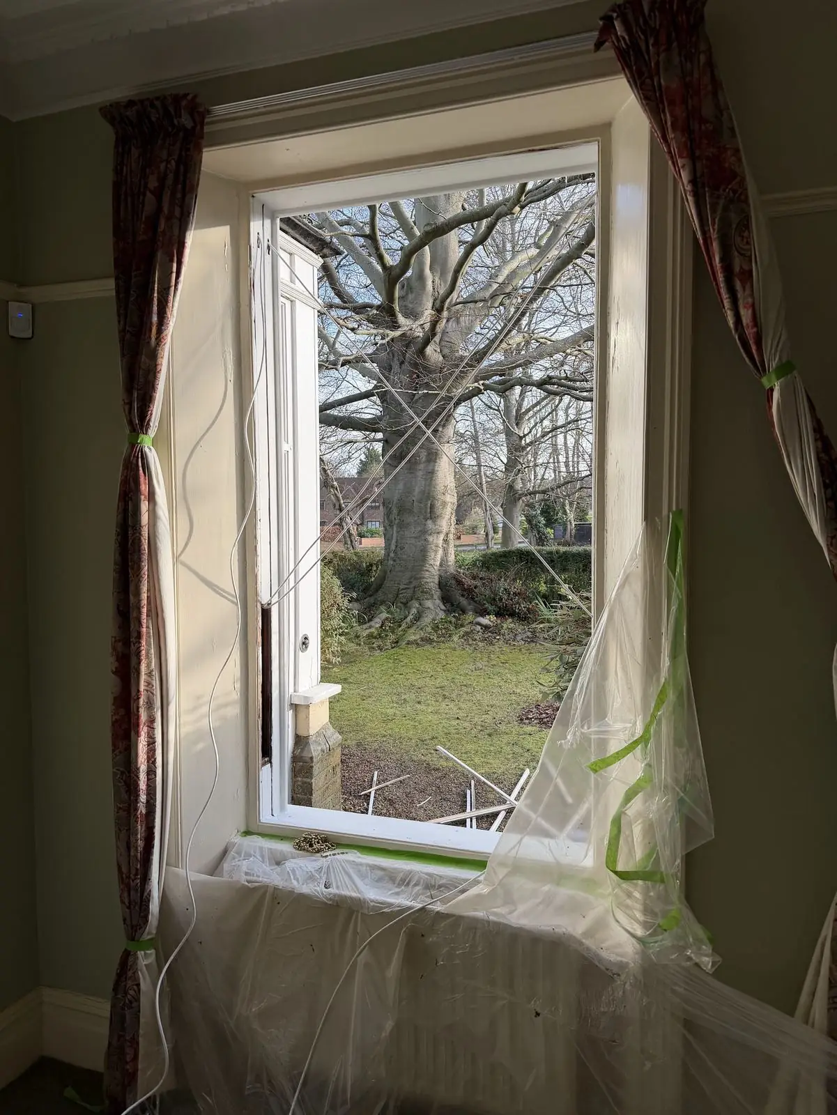Interior view of a sash window mid-cord-change: both sashes are out of the box, dust sheets taped over the cill and radiator, curtains tied back with masking tape, a length of new cord hanging from the pulley pocket.