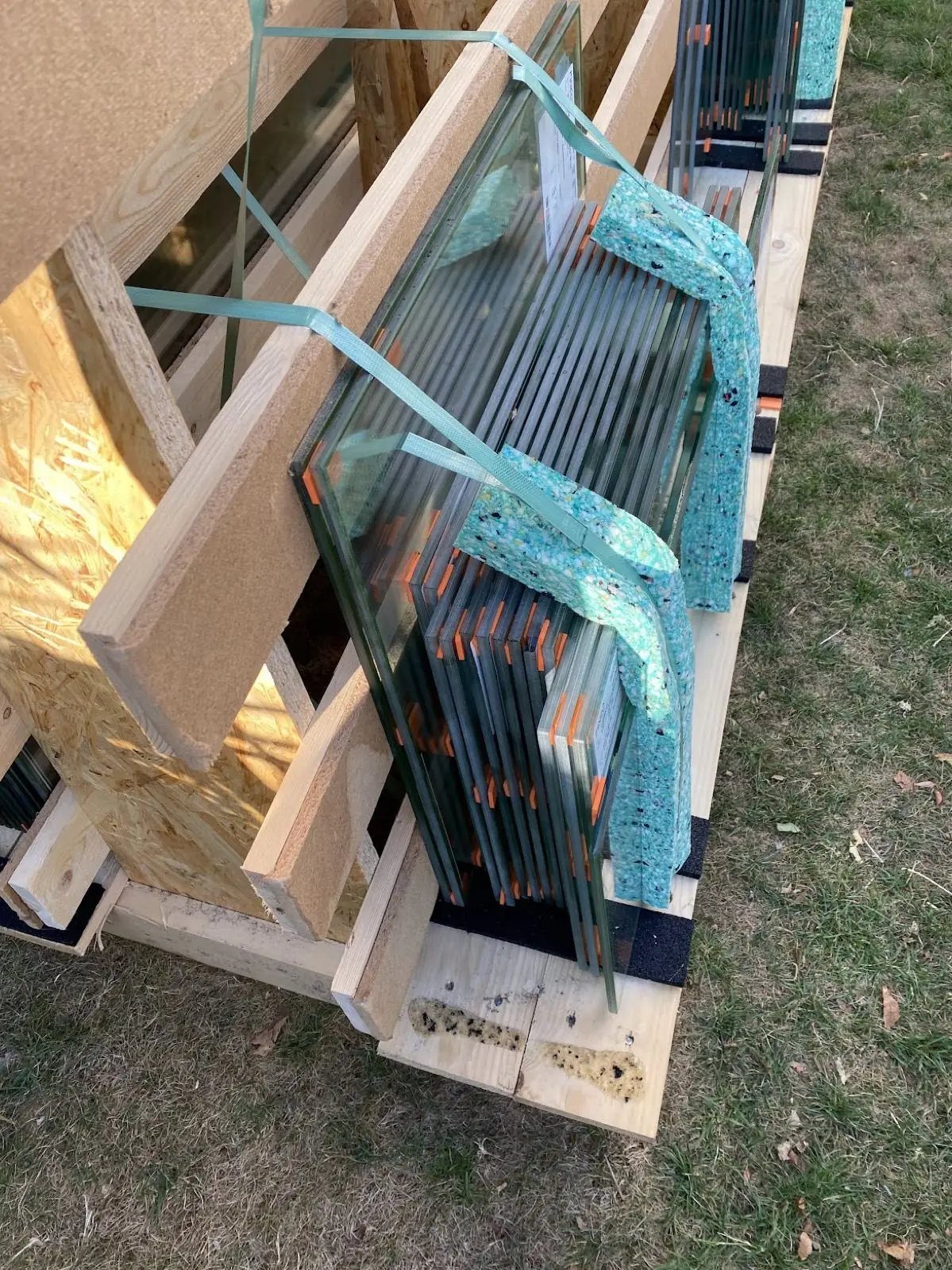 A delivery crate of vacuum-insulating glass units, edge-on, strapped with turquoise webbing — sealed-edge dual-pane construction visible