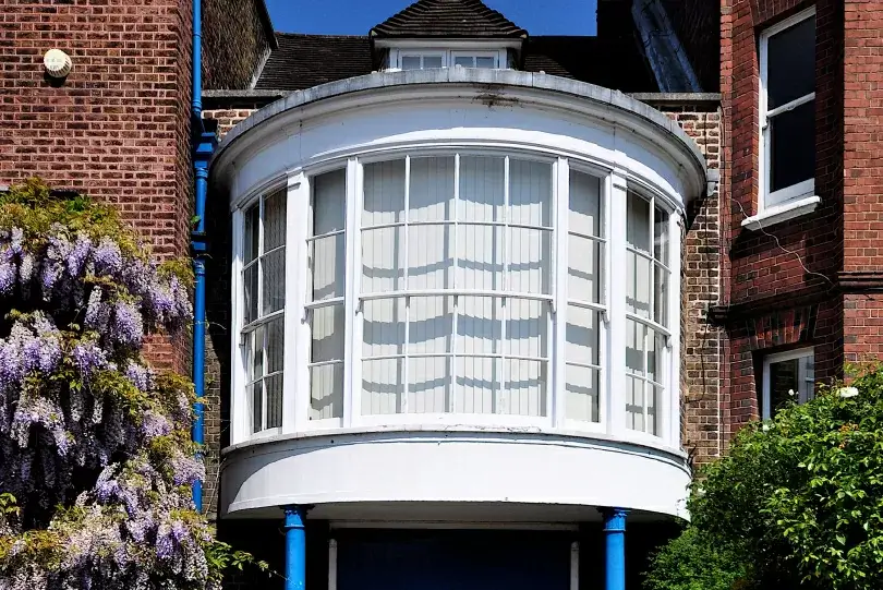 A large bay window with white blinds, reminiscent of the history of sash window styles, is set in a brick building facade, surrounded by purple wisteria on the left.