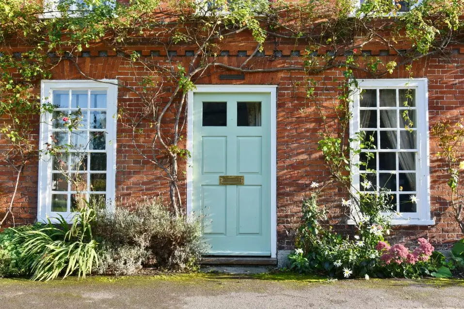 The red brick house facade features a light blue door, flanked by two white-framed sash windows reflecting the history of sash window styles. Climbing plants and greenery gracefully surround the entrance.