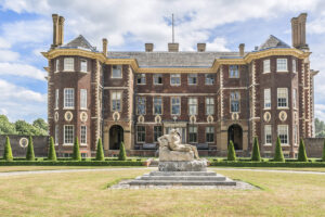 A large historic brick mansion with circular sash windows and tall chimneys, featuring a lawn with a stone sculpture in the foreground.