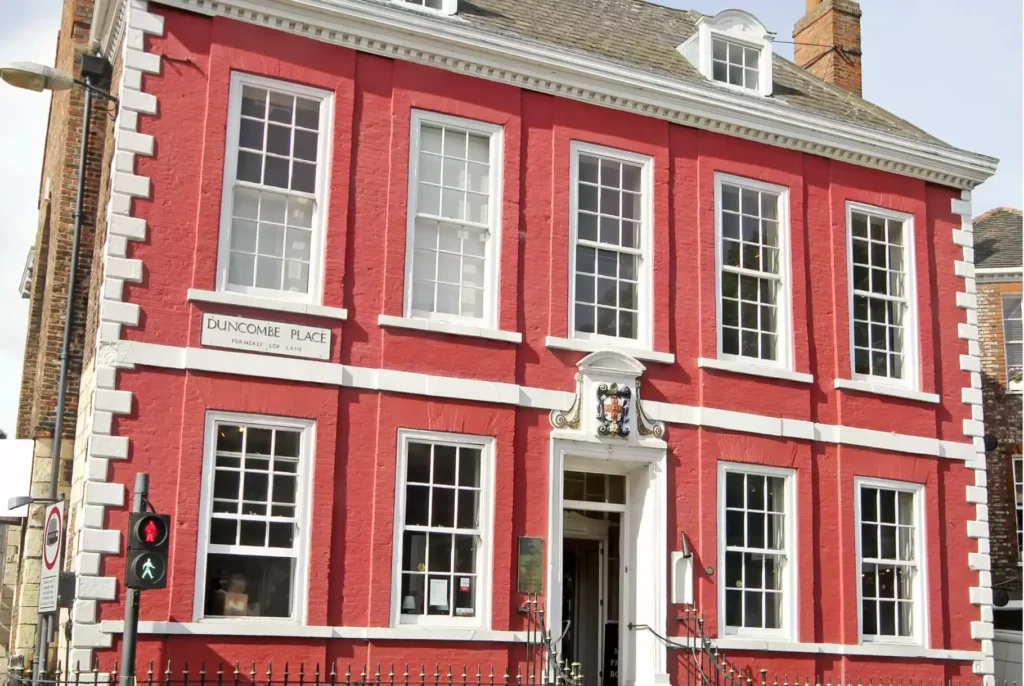 Three-story red brick building with white-trimmed sash windows, showcasing the history of sash window styles, located at Duncombe Place. A coat of arms above the entrance and a pedestrian crossing signal are visible.