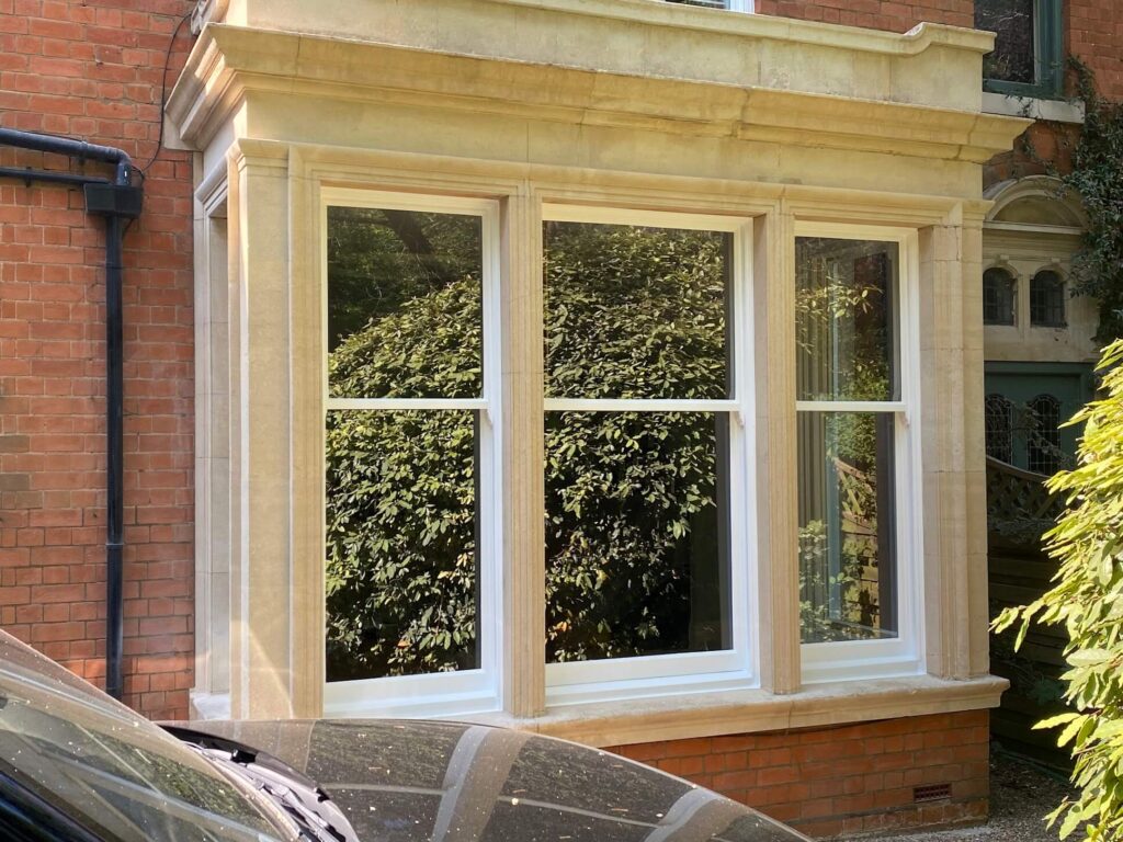 A bay window with three sections and white frames, expertly reflecting the greenery outside, hints at the sash window restoration craftsmanship found in Northamptonshire’s charming brick buildings.