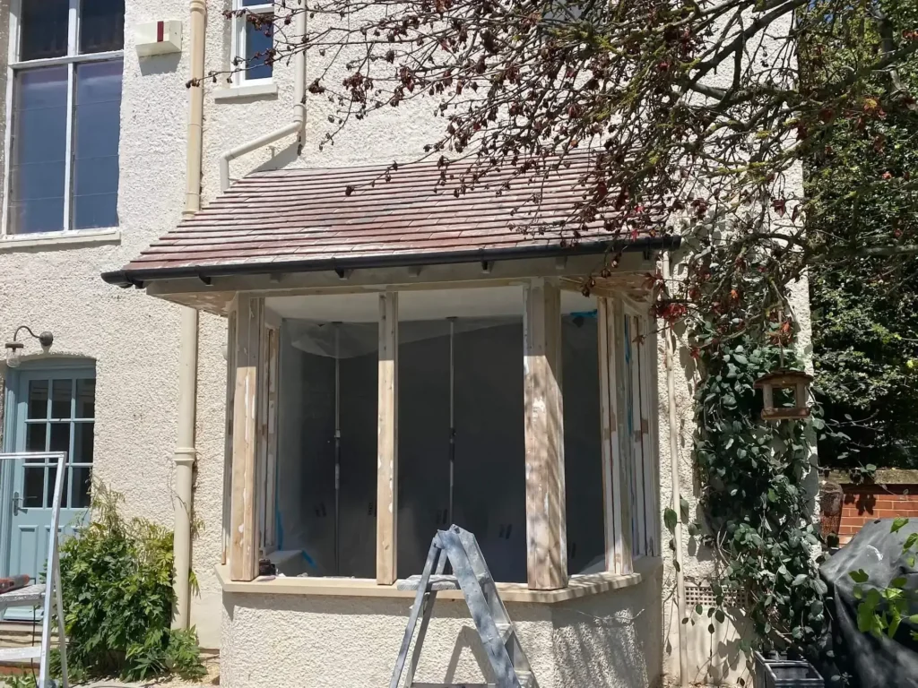 A sash window restoration in Northamptonshire reveals exposed wooden frames on a cream-colored house. A stepladder stands ready while a tree with sparse leaves casts intriguing shadows on the wall.