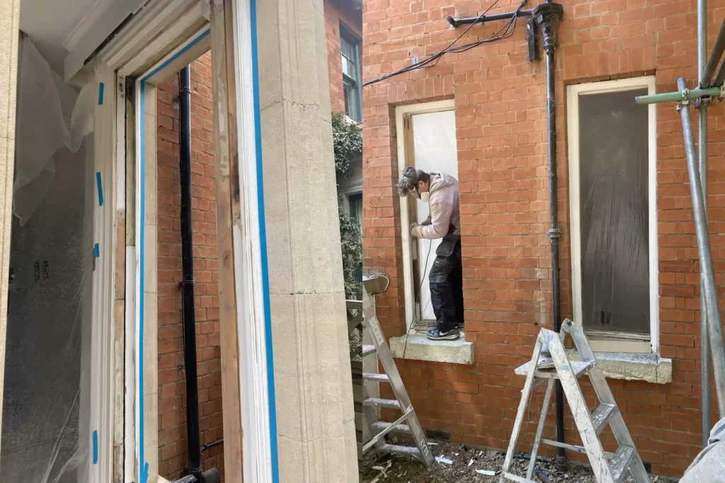 A person stands on a window ledge of a brick building, conducting sash window restoration in Northamptonshire while balancing on a ladder. Multiple ladders are positioned nearby.