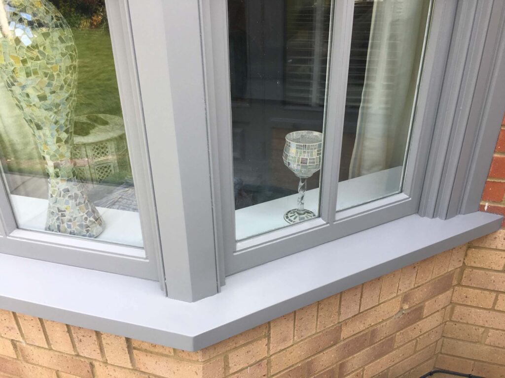 A gray-framed window with Georgian glazing bars showcases mosaic-patterned glass items on the sill: a vase and a wine glass. The window overlooks a serene garden area, blending tradition with modern evolution.