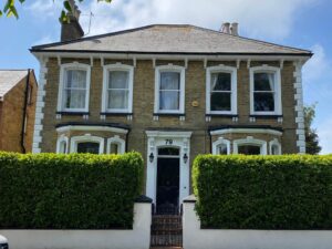 Two-story brick house with a dark door numbered , featuring elegant Victorian sash windows as a timeless feature, and framed by a tall green hedge.