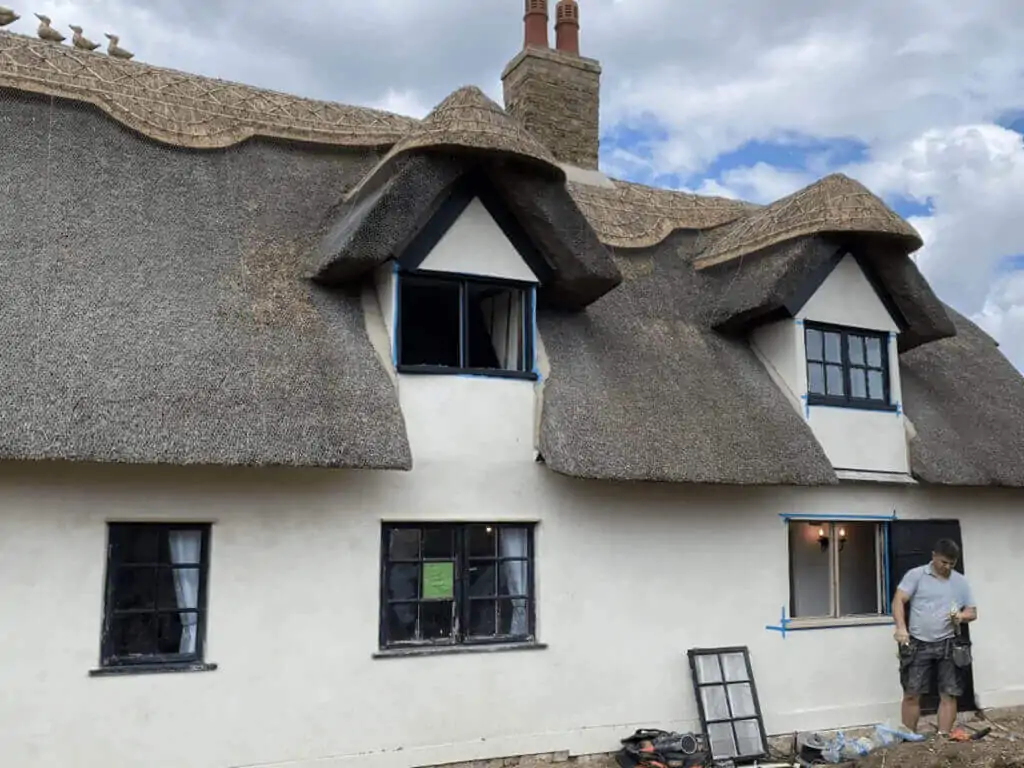 A person stands outside a charming white thatched cottage, with windows taped for painting. Tools are scattered on the ground, hinting at an upgrade to double-glazed, cottage-style windows.