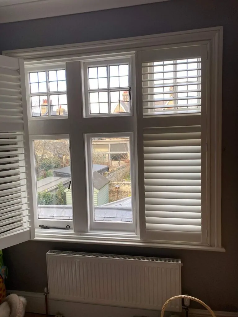 The white window shutters are partially open, revealing a view of a garden shed and neighboring buildings. A radiator sits below the window, where natural light streams in. You might wonder, can you install secondary glazing with shutters?.