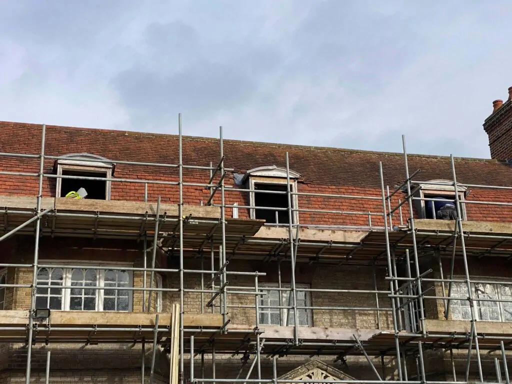 A building under construction stands with scaffolding, where workers can be glimpsed through openings near the roof. The overcast sky looms above as efforts to restore wooden windows proceed diligently.