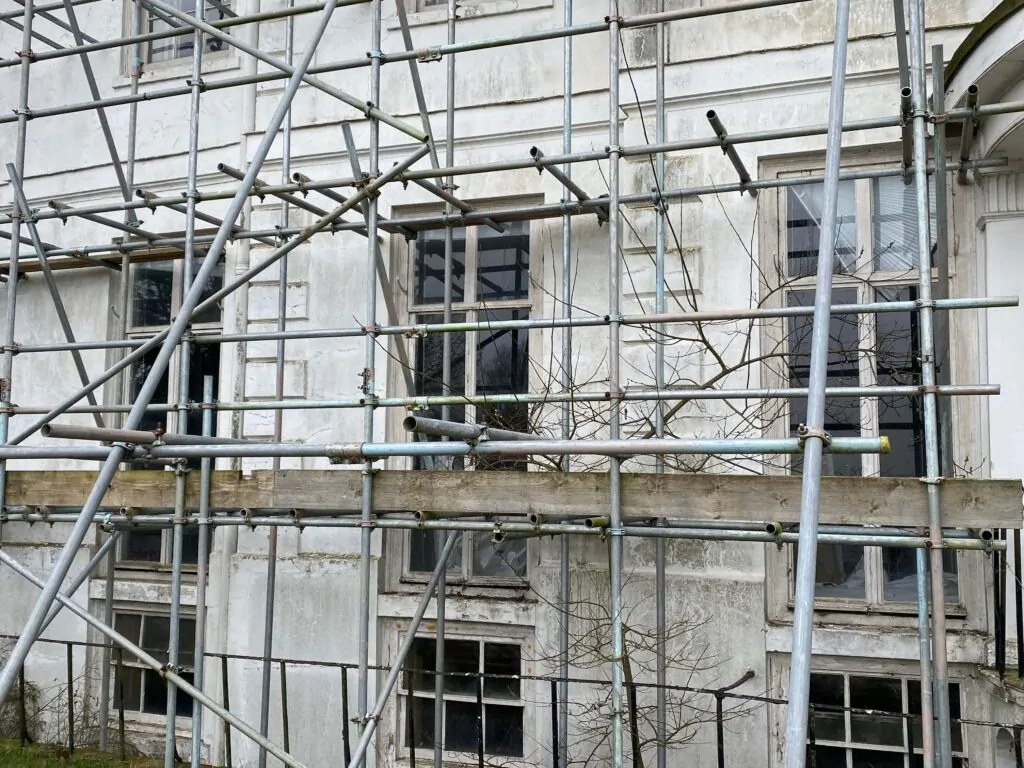 Scaffolding stands in front of the old, weathered building with multiple windows, likely for maintenance or to restore wooden windows. Sparse branches are visible in front of the structure.